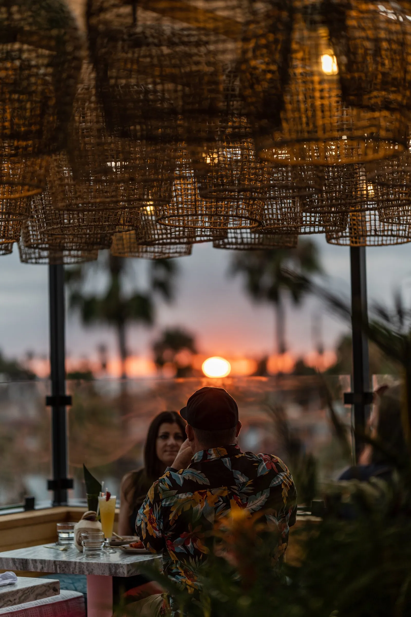 Sunset viewed from Cococabana rooftop through glass railing, palms silhouetted against orange sky